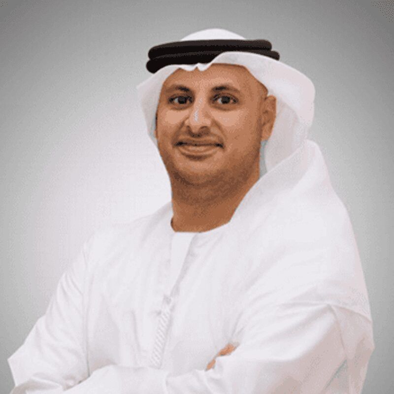 Confident Middle Eastern man in traditional Emirati attire, including white kandura and black agal, standing with arms crossed and smiling against a neutral background