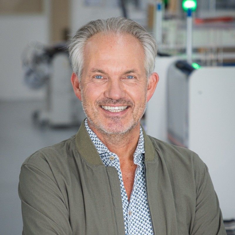 Smiling middle-aged man with gray hair wearing a green jacket, standing in a modern workspace or lab environment.