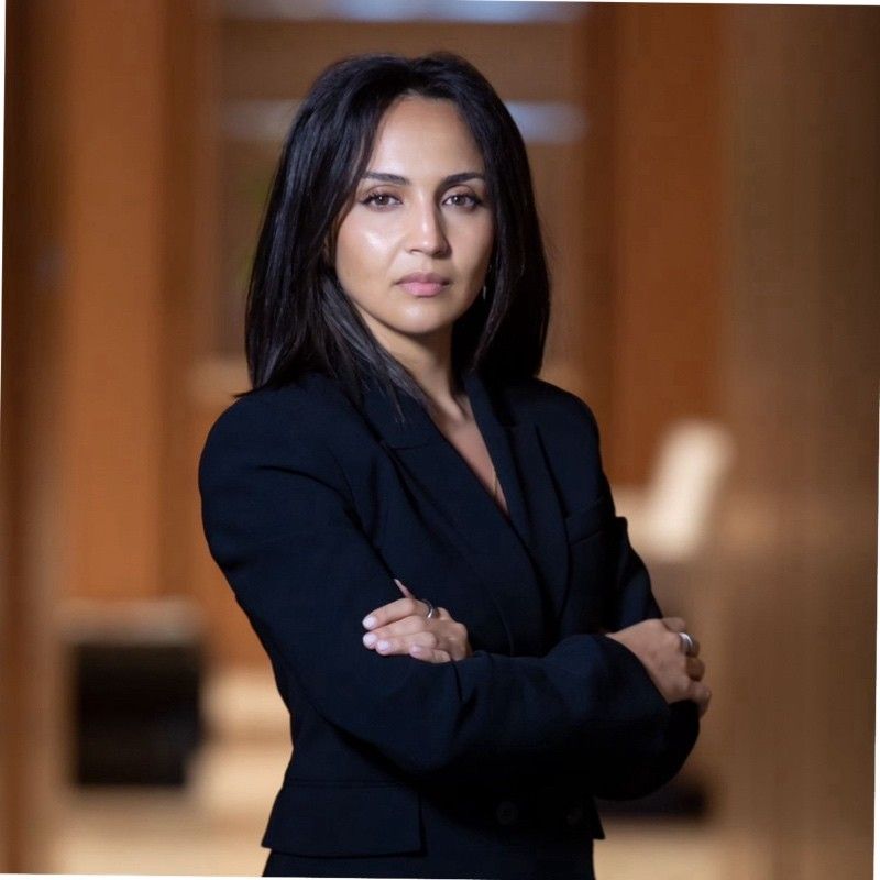 Confident woman in a black blazer standing with arms crossed in a warmly lit modern hallway.