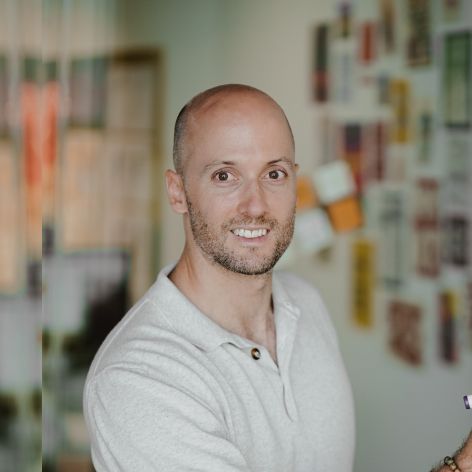 Smiling man in a light grey polo shirt standing in a creative office space with colorful notes and materials pinned on the wall behind him