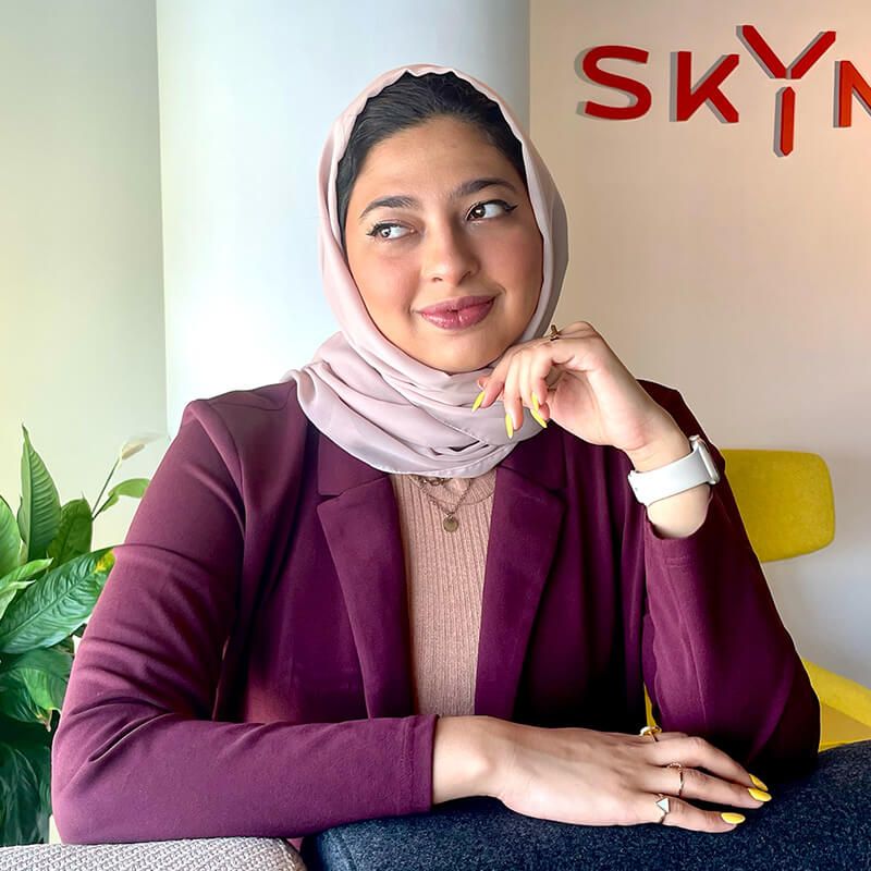Smiling woman in maroon blazer and pink hijab sitting near SKYNE branding