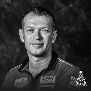 Black and white portrait of a Dakar Rally participant wearing a branded team shirt with sponsor logos, against a dark studio background.