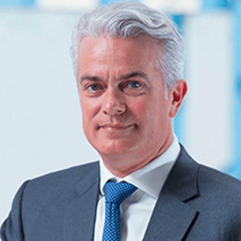 Portrait of a mature man with gray hair in a navy suit and blue tie, posing confidently in a corporate setting.