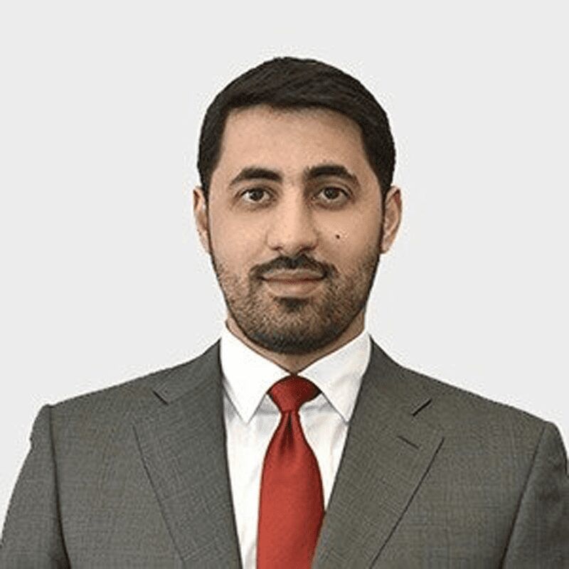 Professional man with short dark hair and trimmed beard wearing a gray suit, white shirt, and red tie, posing against a plain light background