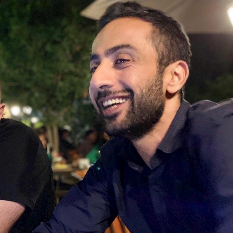 Smiling man in a dark shirt sitting outdoors at night, with blurred greenery and ambient lights in the background.