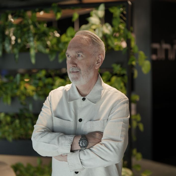 Elderly man with gray hair and beard focused on sketching or writing at a desk, wearing a light-colored buttoned shirt in a softly lit indoor setting