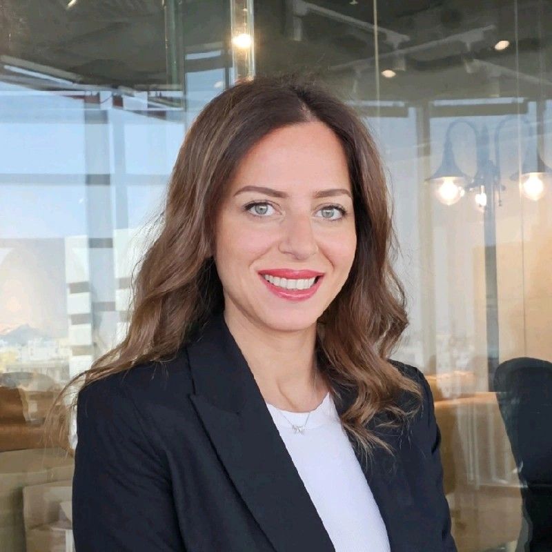 Professional woman with long brown hair wearing a black blazer and white top, smiling in a modern glass-walled office setting