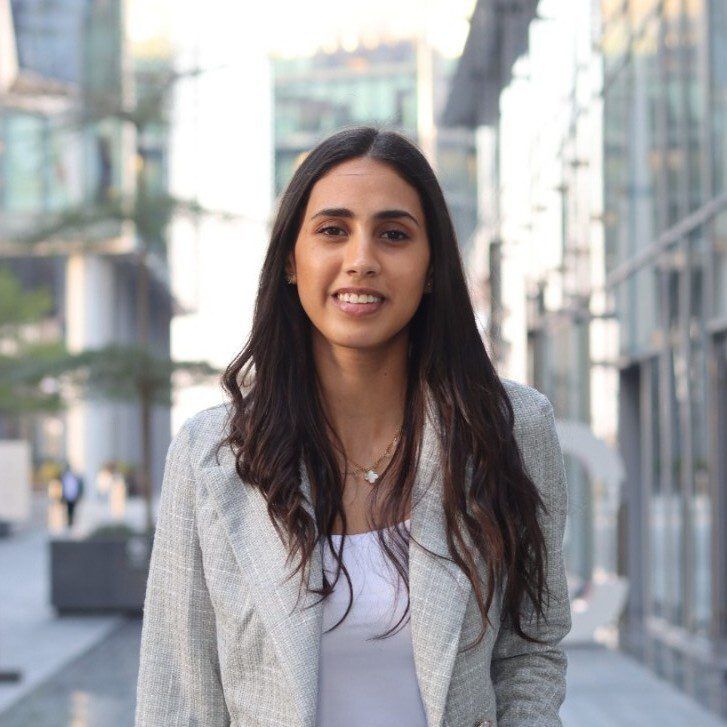 Young professional woman with long dark hair wearing a light blazer and white top, standing confidently in a modern urban business district