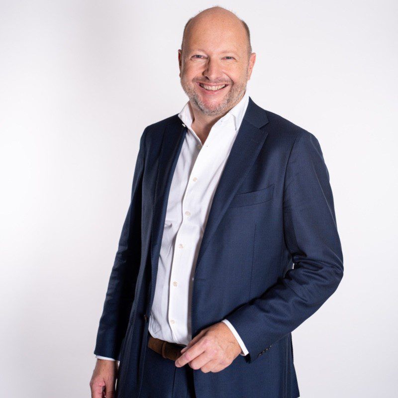 Smiling man in a navy blue suit with a white shirt standing against a plain light background