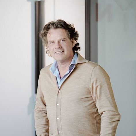 Smiling man with wavy brown hair wearing a beige cardigan and light blue shirt, standing in a bright modern office corridor