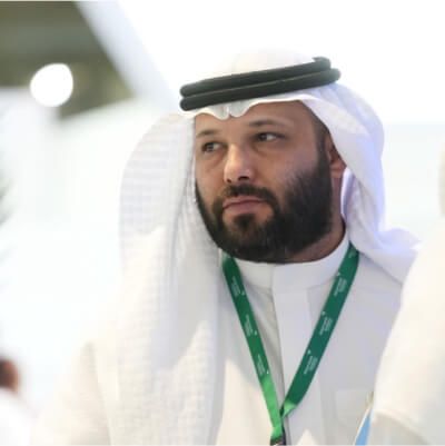 Arab man in traditional white thobe and black agal, wearing a green lanyard badge, looking thoughtful at an event