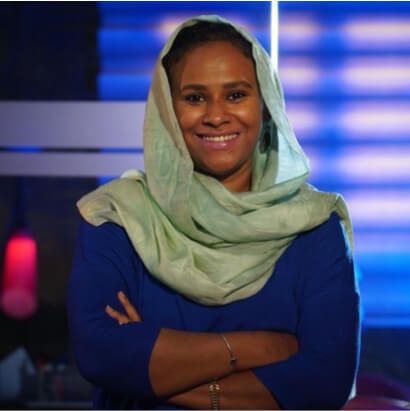 Confident woman in green headscarf and blue top smiling with arms crossed in a modern studio background