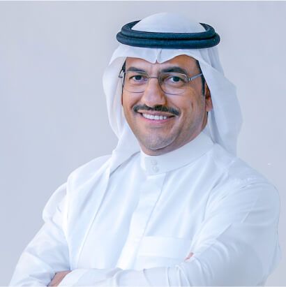 Smiling Arab man in traditional white thobe and headscarf with glasses, arms crossed against light background.