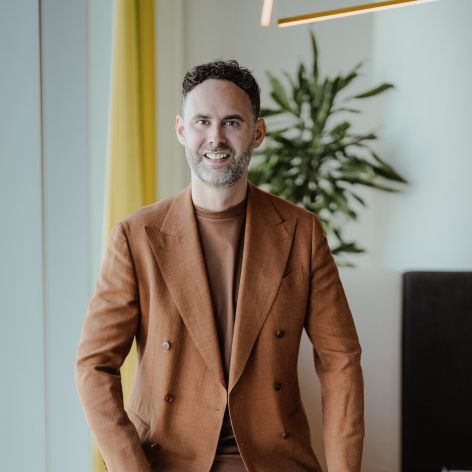 Smiling man in a brown suit standing indoors near a plant and modern lighting
