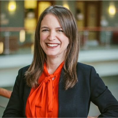 Smiling professional woman with straight brown hair wearing a black blazer and vibrant orange blouse, standing in a modern office atrium