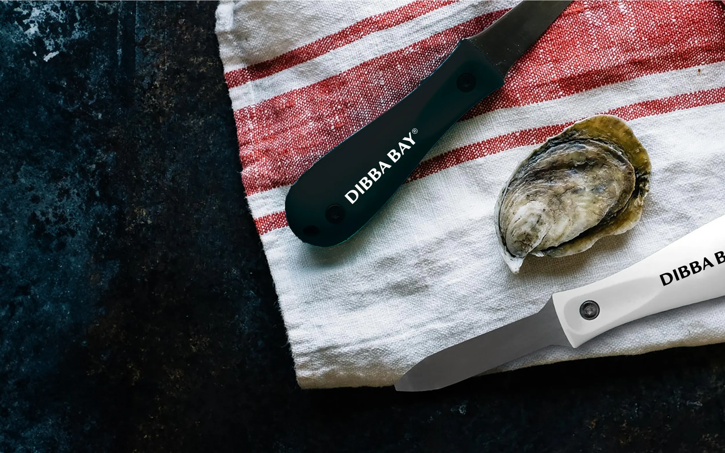 Dibba Bay branded oyster knives placed on a striped cloth next to a fresh oyster, styled for premium seafood branding