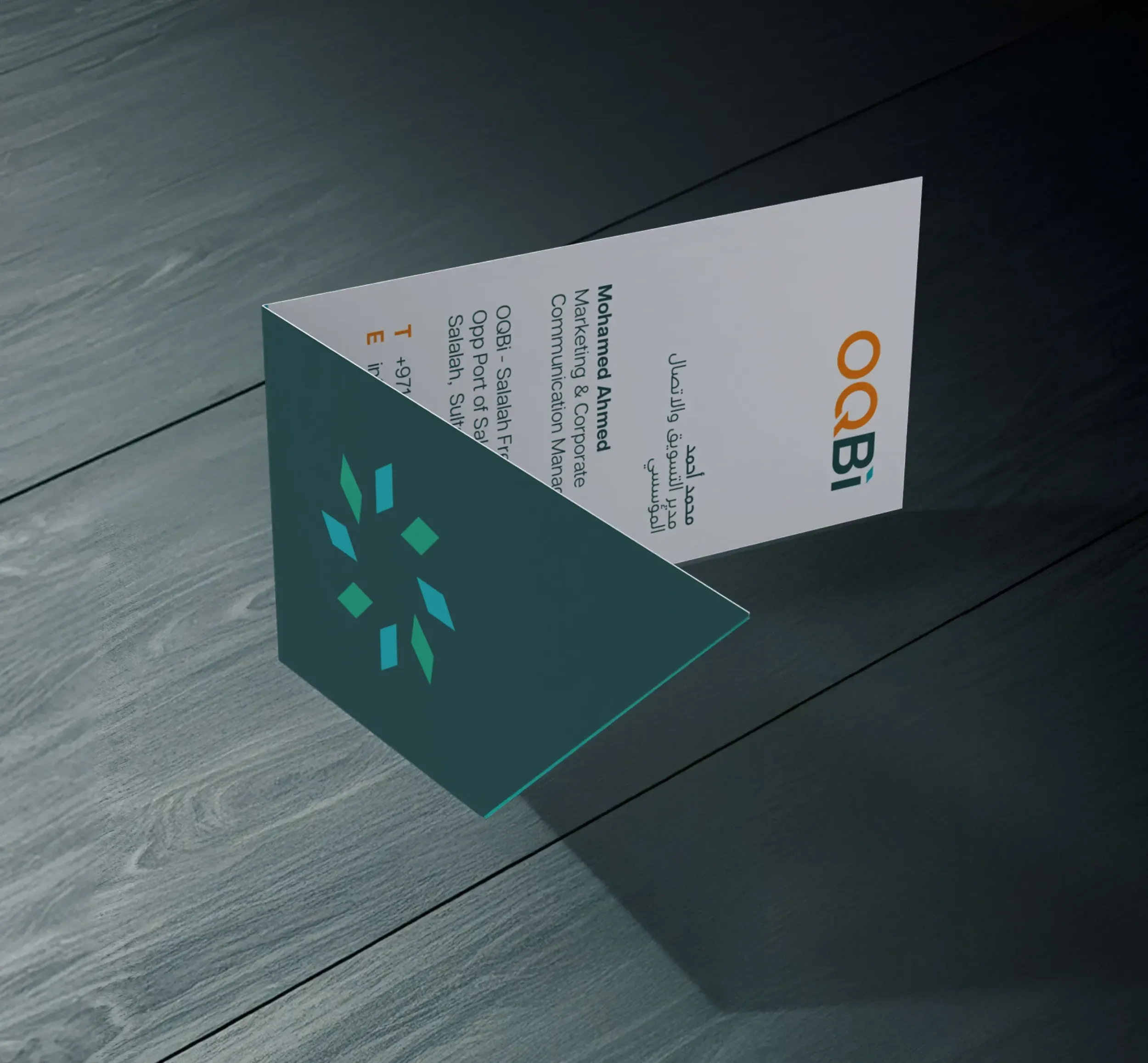 Modern vertical business card design for OQBI, displayed standing on a dark wooden surface with bilingual English-Arabic contact details and geometric teal logo