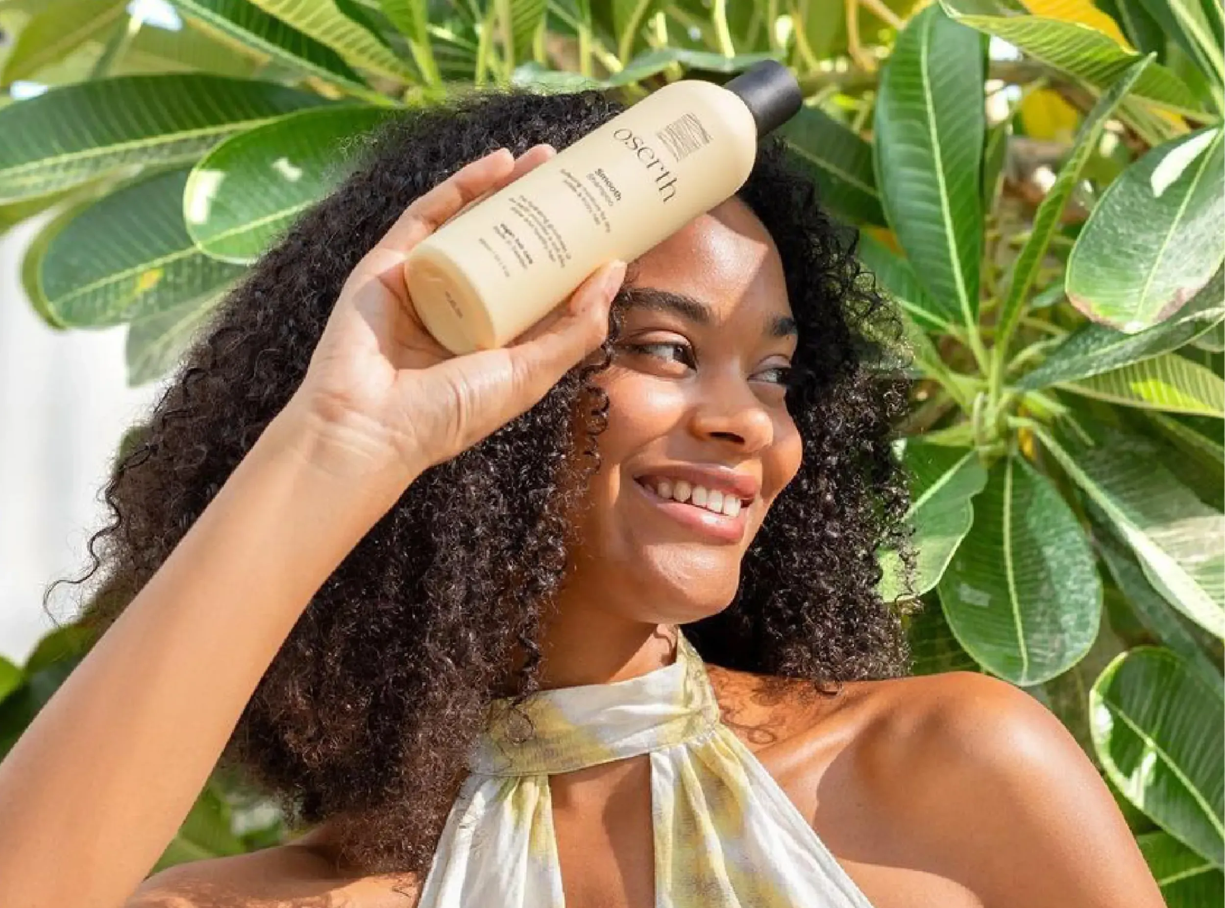Smiling woman with curly hair holding Oserth shampoo bottle against green leaves, conveying natural beauty and product connection to nature, reflecting Skyne’s lifestyle branding.