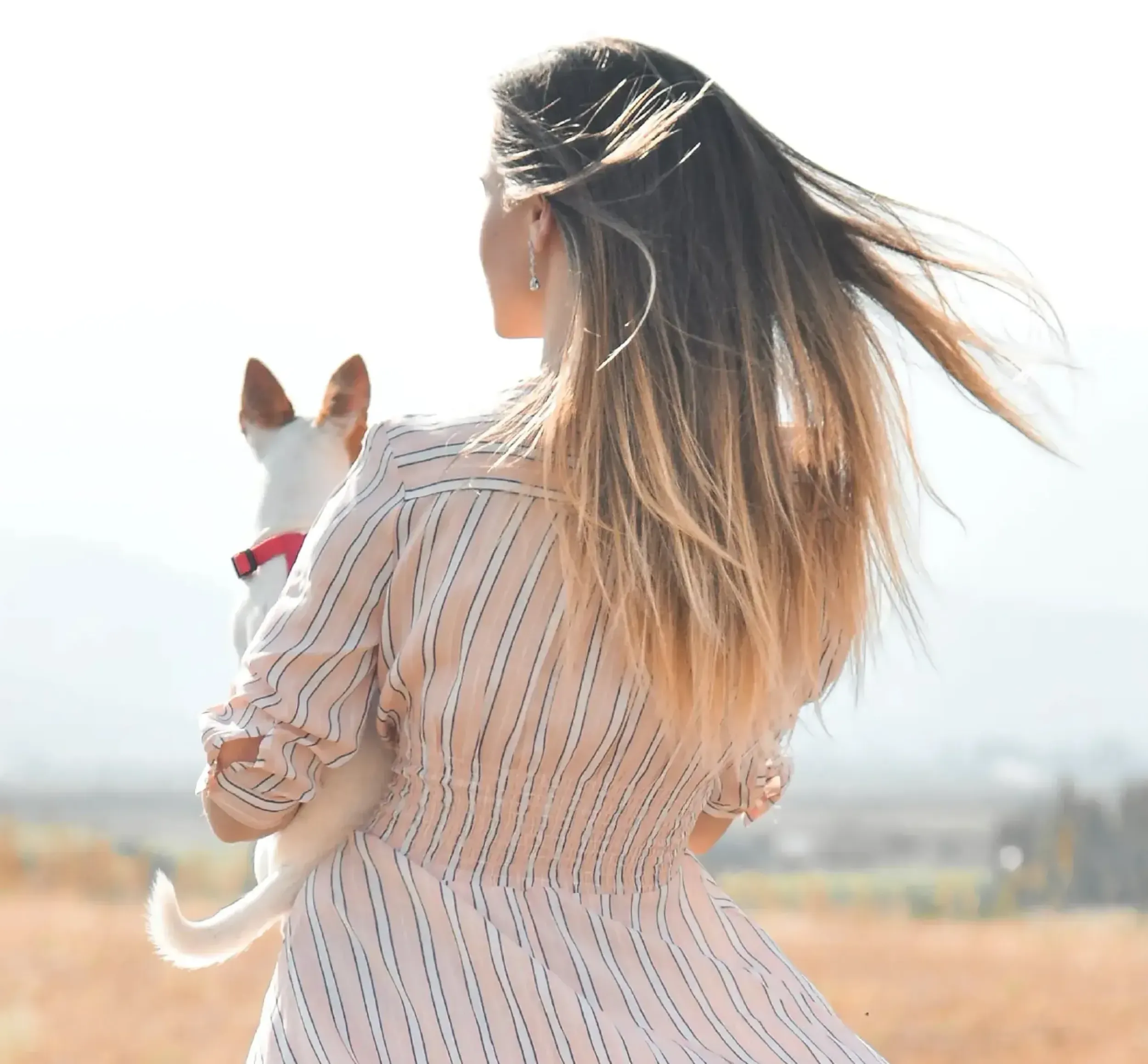 Woman holding a small white dog with hair blowing in the wind, evoking freedom and natural beauty, aligning with Oserth’s brand message and Skyne’s lifestyle branding.