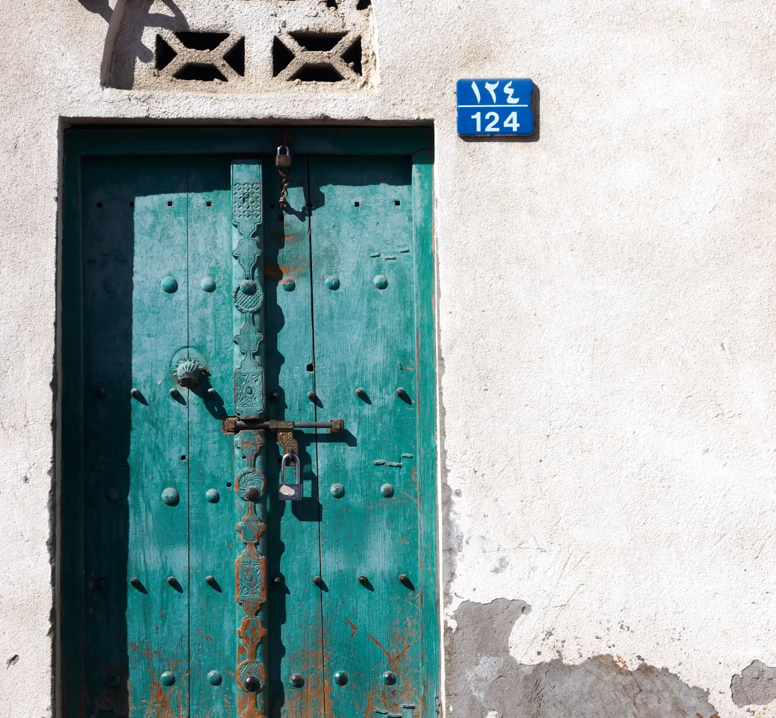 Rustic weathered teal wooden door with ornate metalwork and padlock, set against a white textured wall with number "124" plaque. 