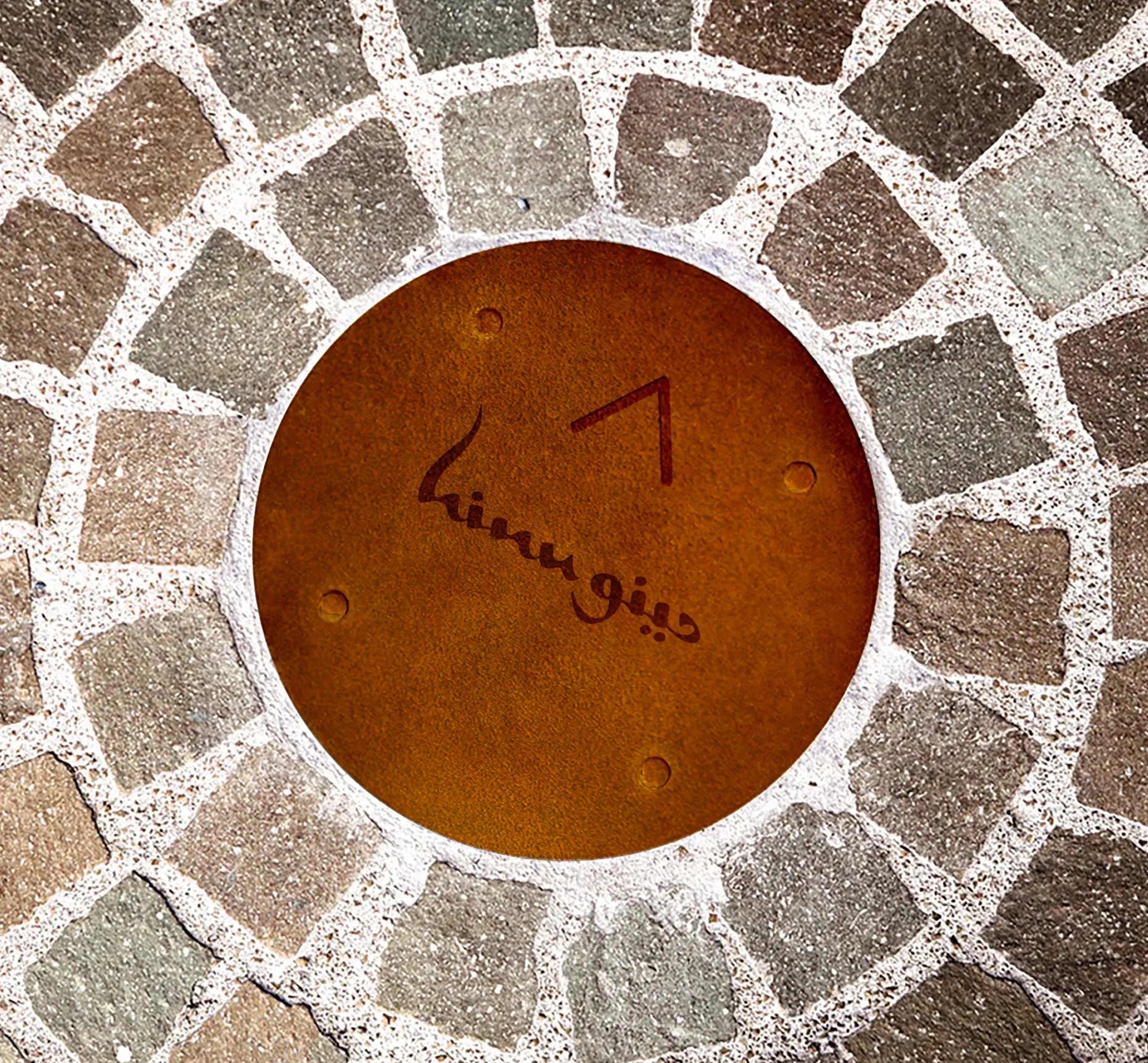 Circular rustic metal plaque with Hinu Bay logo and an arrow embedded in a cobblestone pathn. Skyne creative agency delivers comprehensive branding and environmental design for destinations in Dubai, Riyadh, and Jeddah.