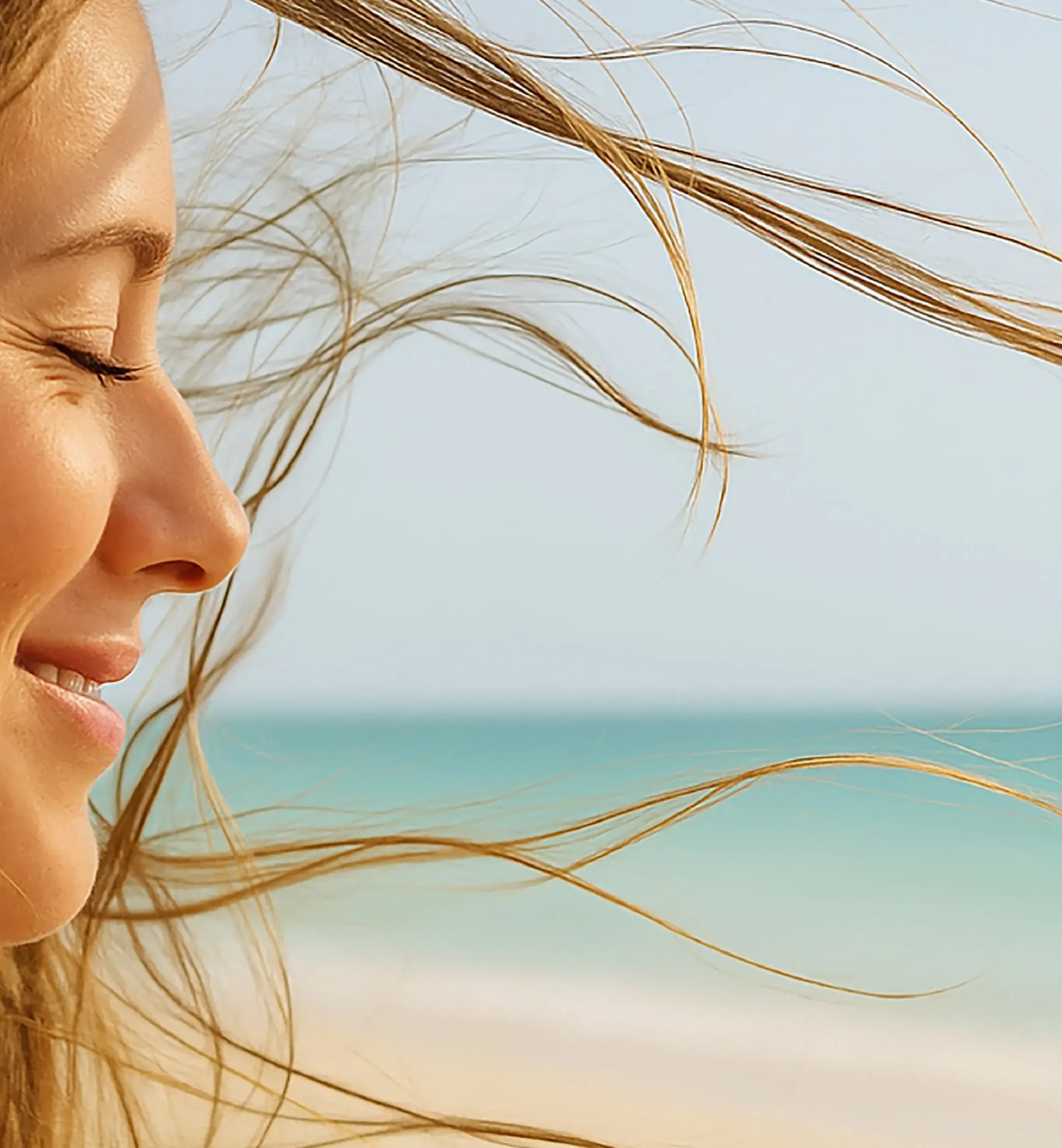 Profile of a smiling woman with eyes closed, hair blowing in the wind, with a serene beach and turquoise sea in the background. This image reflects the branding efforts of Skyne, a creative agency specializing in branding in Dubai, Riyadh, and Jeddah.