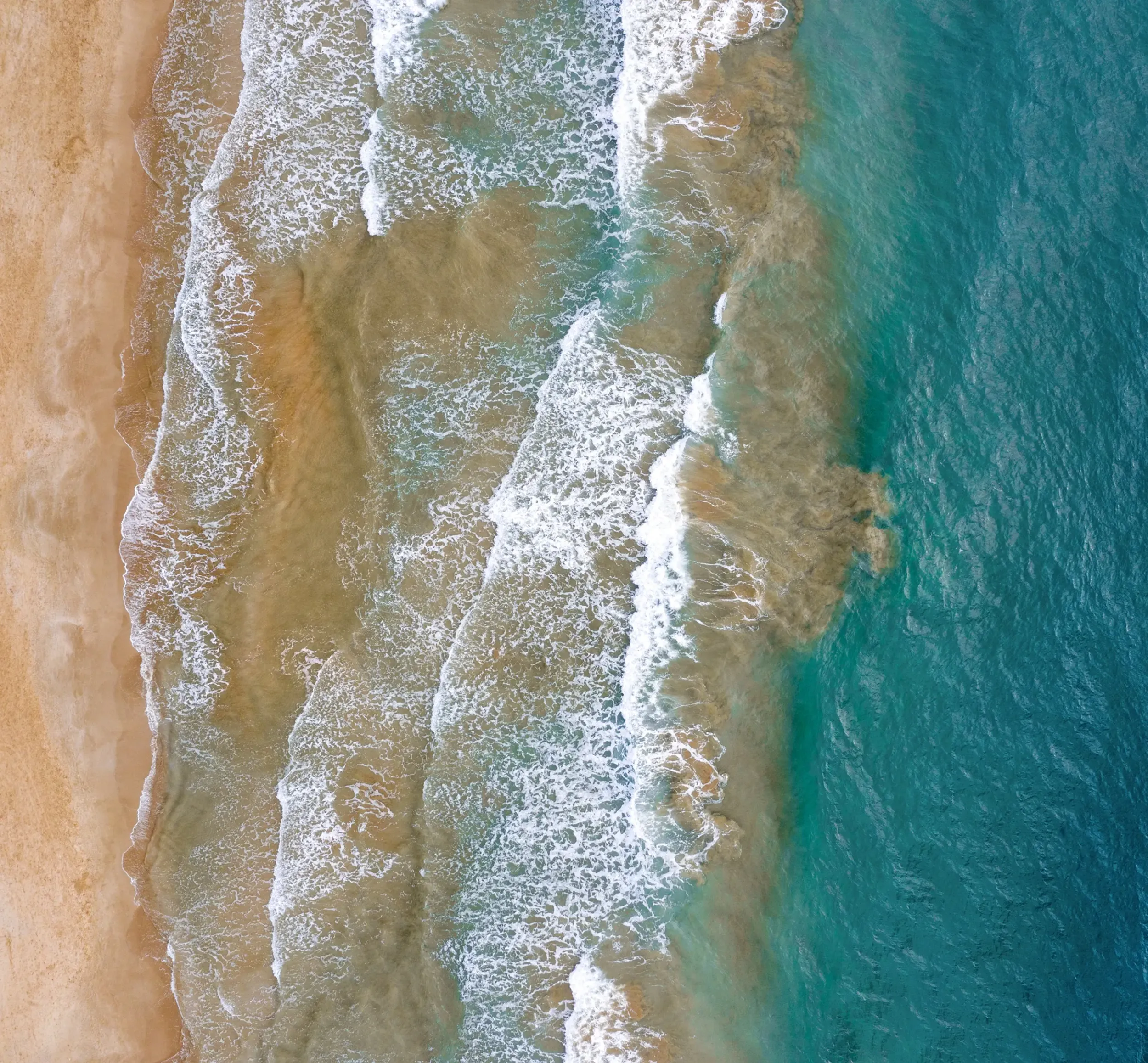 Aerial view of ocean waves crashing onto a sandy beach, showcasing clear blue water and textured shoreline patterns