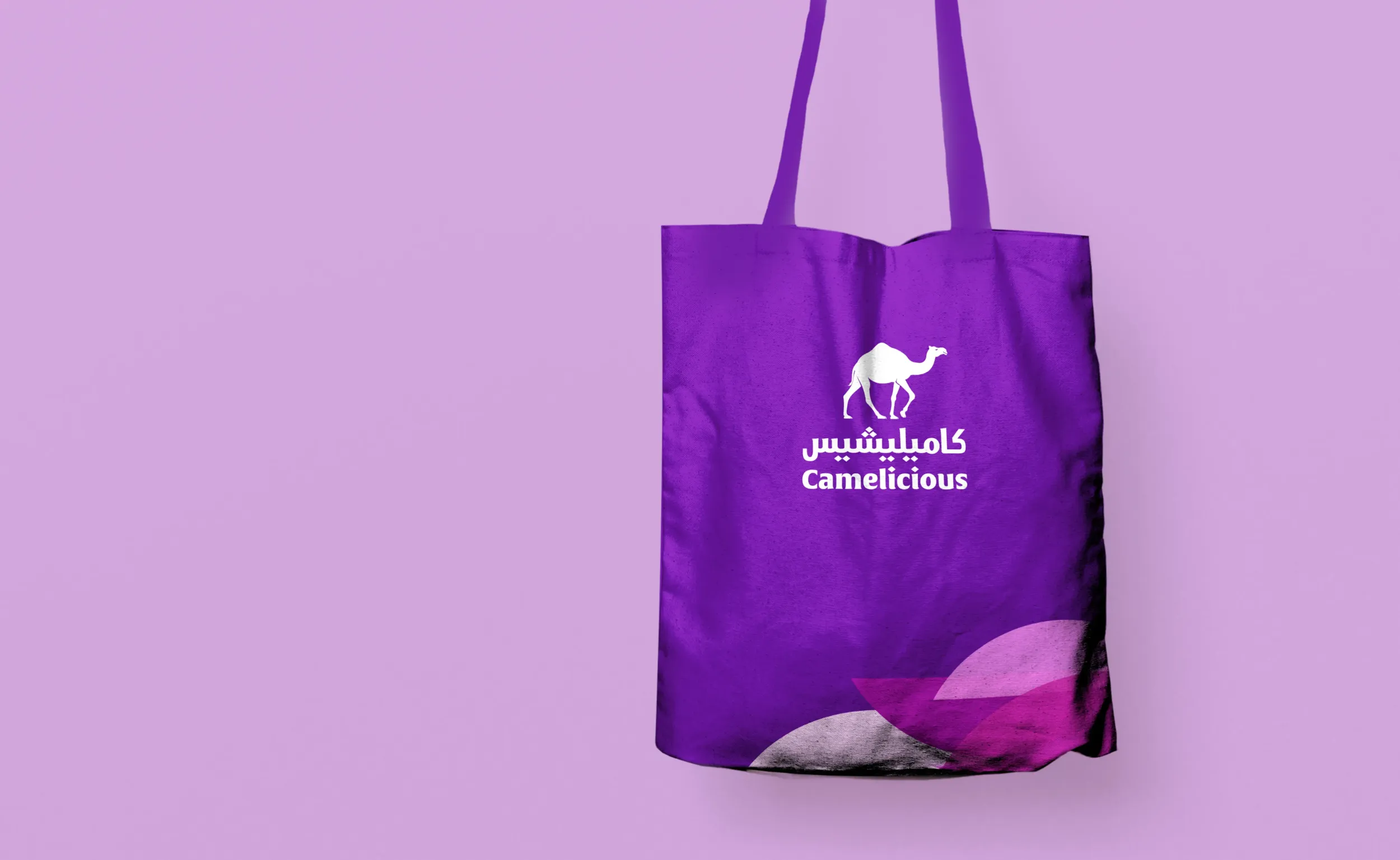 Purple tote bag with the Camalicious logo featuring a white camel icon and bilingual Arabic-English text, placed against a lavender background